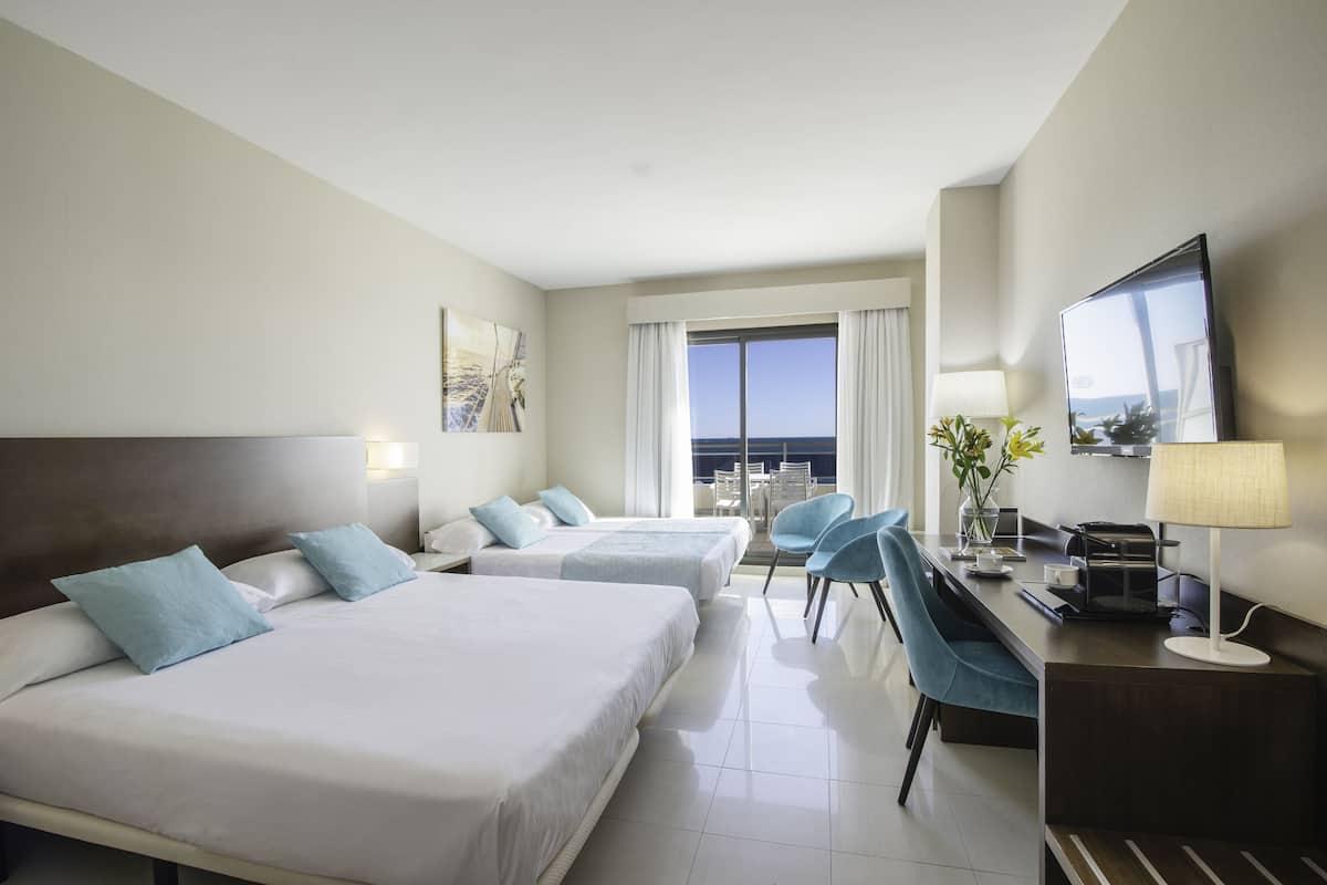 A hotel room with two beds, a desk with a lamp, a chair, and a large window with a view.