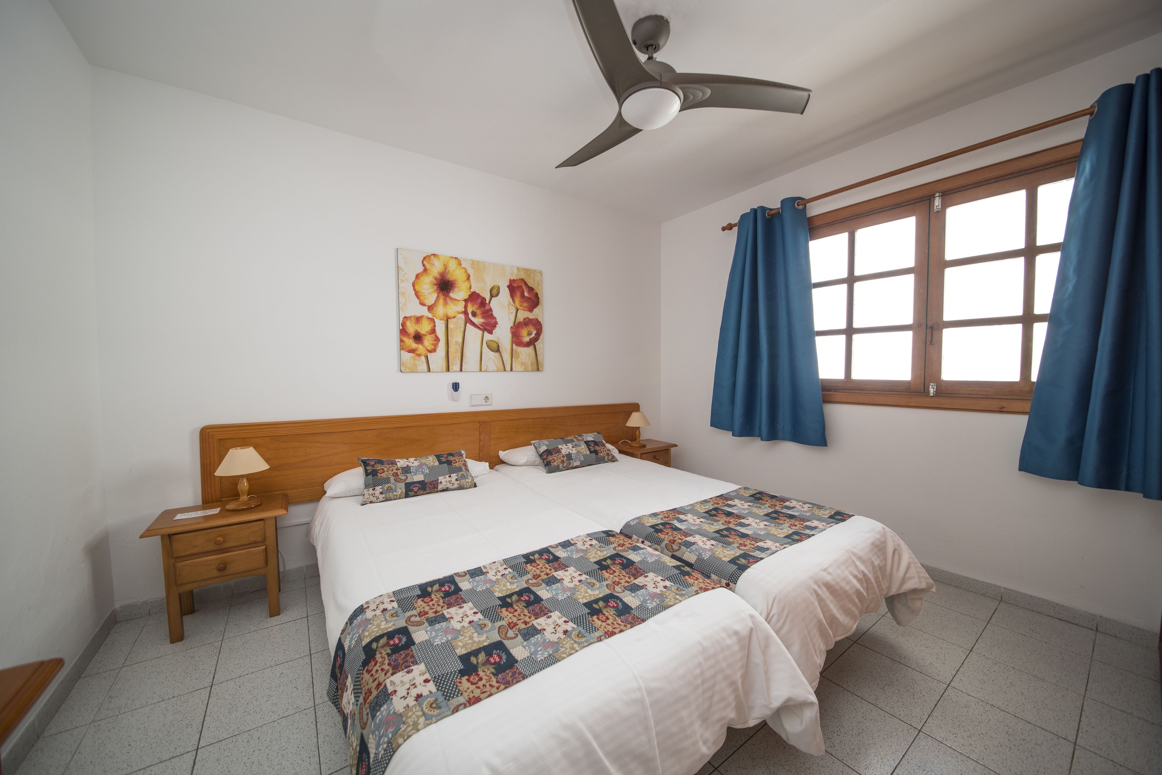 Une chambre à coucher avec un lit, une table de chevet, un ventilateur de plafond, une fenêtre avec des rideaux et un tableau accroché au mur.