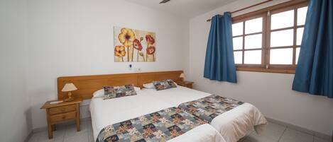 Une chambre à coucher avec un lit, une table de chevet, un ventilateur de plafond, une fenêtre avec des rideaux et un tableau accroché au mur.