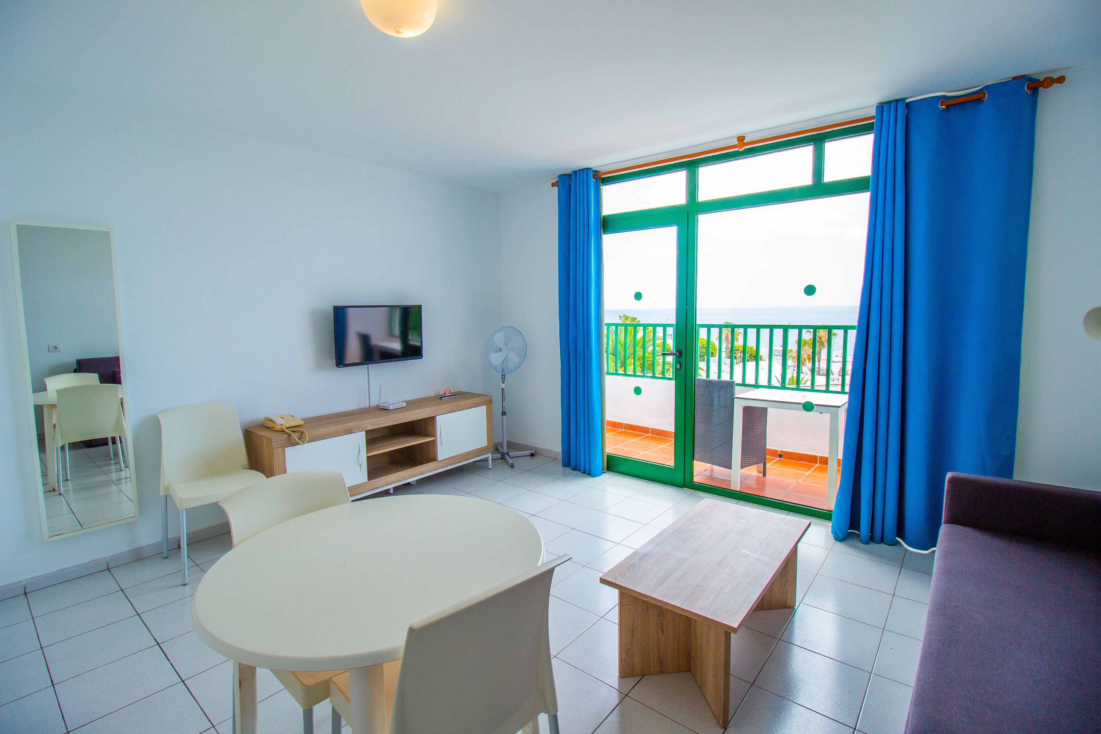 Une pièce comprenant une table à manger, des chaises, un téléviseur, un ventilateur et un balcon avec des rideaux bleus.