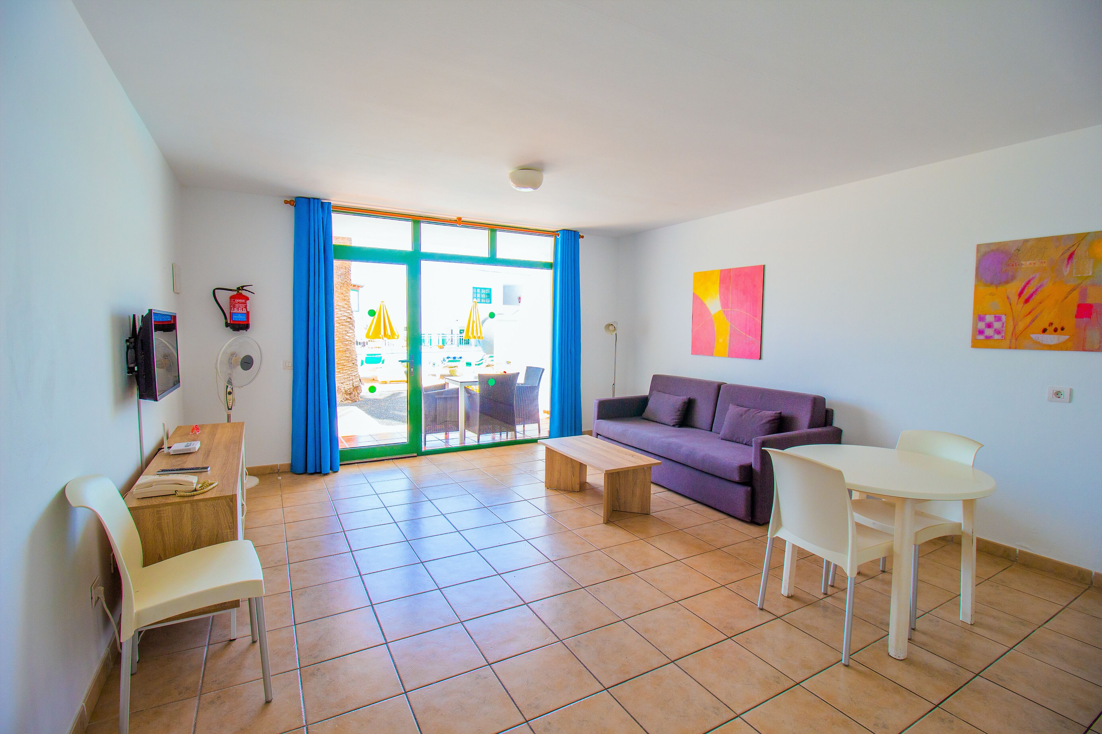 Una sala de estar con sofá, mesa de comedor, sillas, televisor y ventana con cortinas.