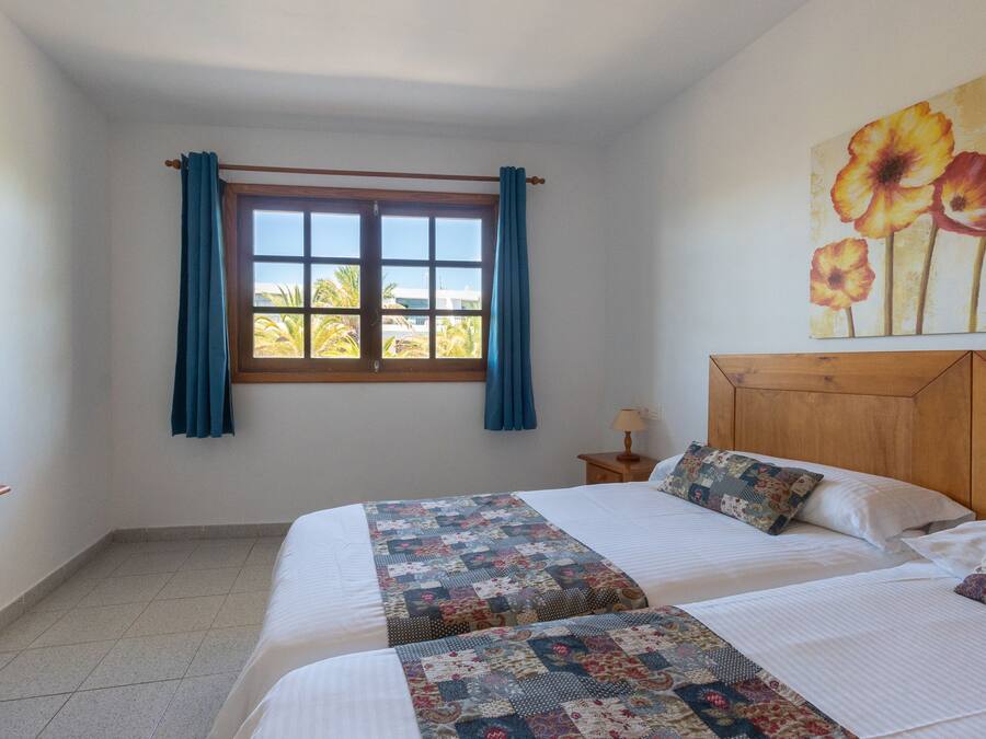 Un dormitorio con dos camas, una ventana con cortinas, un cabecero de madera, una mesita de noche y un cuadro floral en la pared.