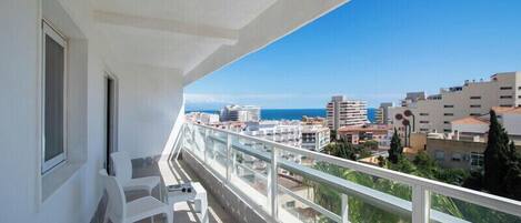 A balcony with white chairs and a glass railing overlooking a cityscape and the sea.