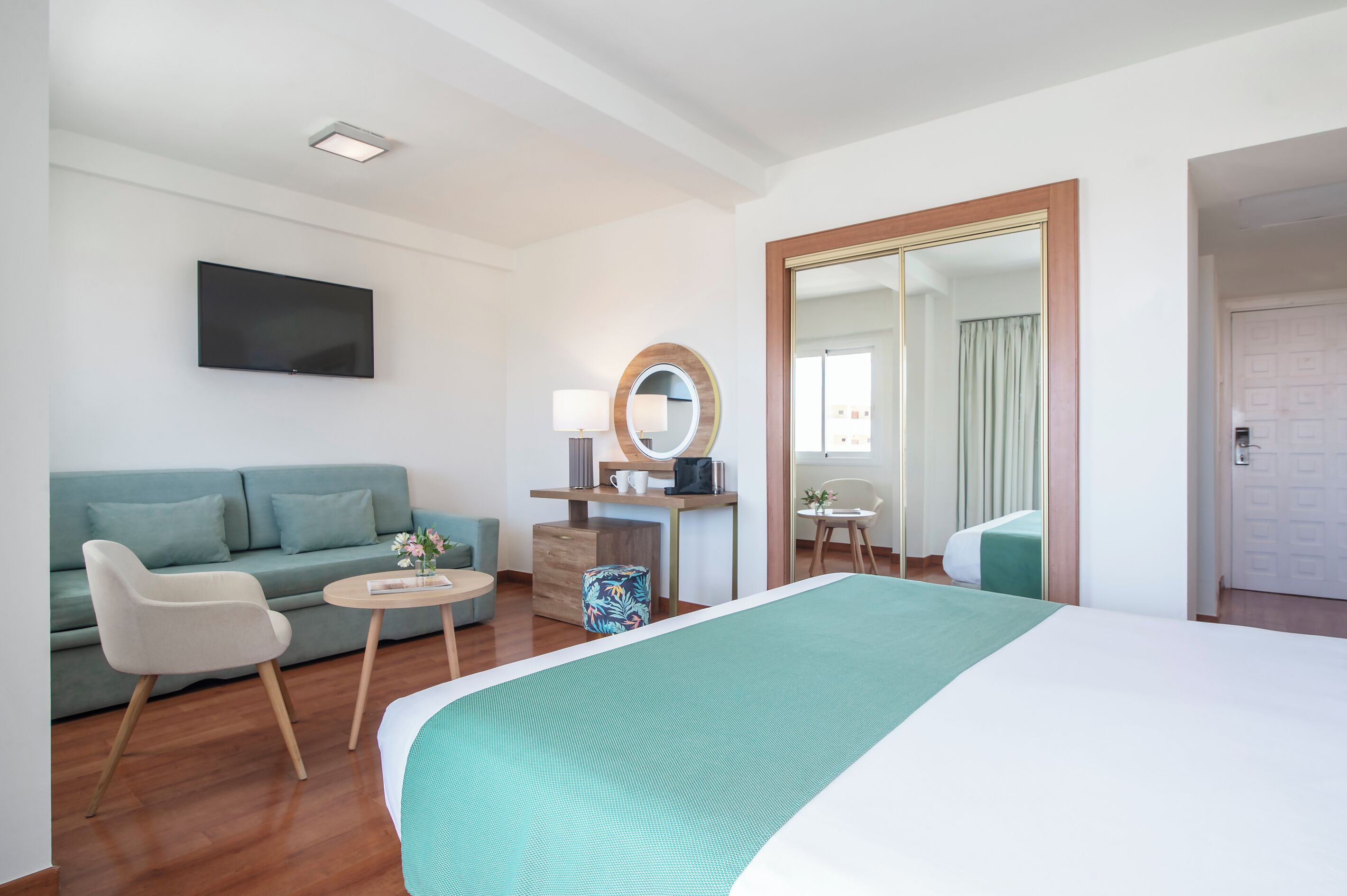 A hotel room with a bed, a sofa, a desk, a chair, a TV, and a mirror.