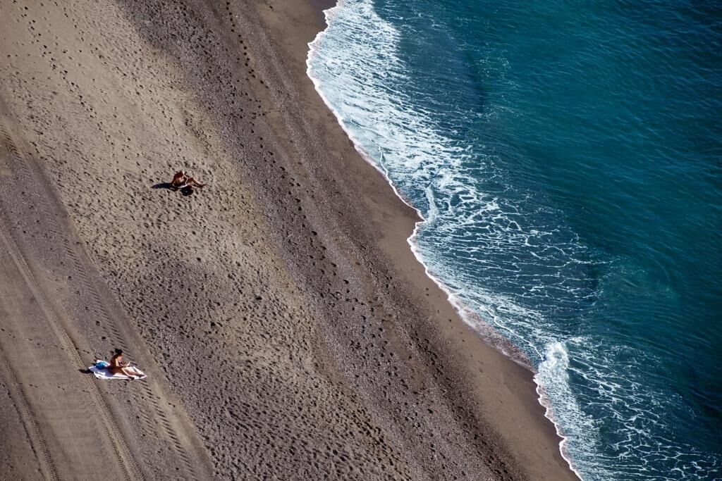 An aerial view of a beach with two people lying on towels, a clear sky, and waves approaching the shore.