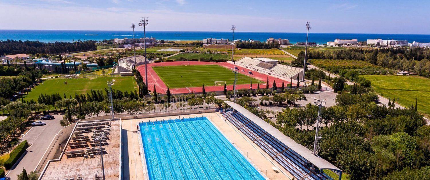 Vista aérea de un complejo deportivo con un estadio grande, una piscina y zonas verdes circundantes.
