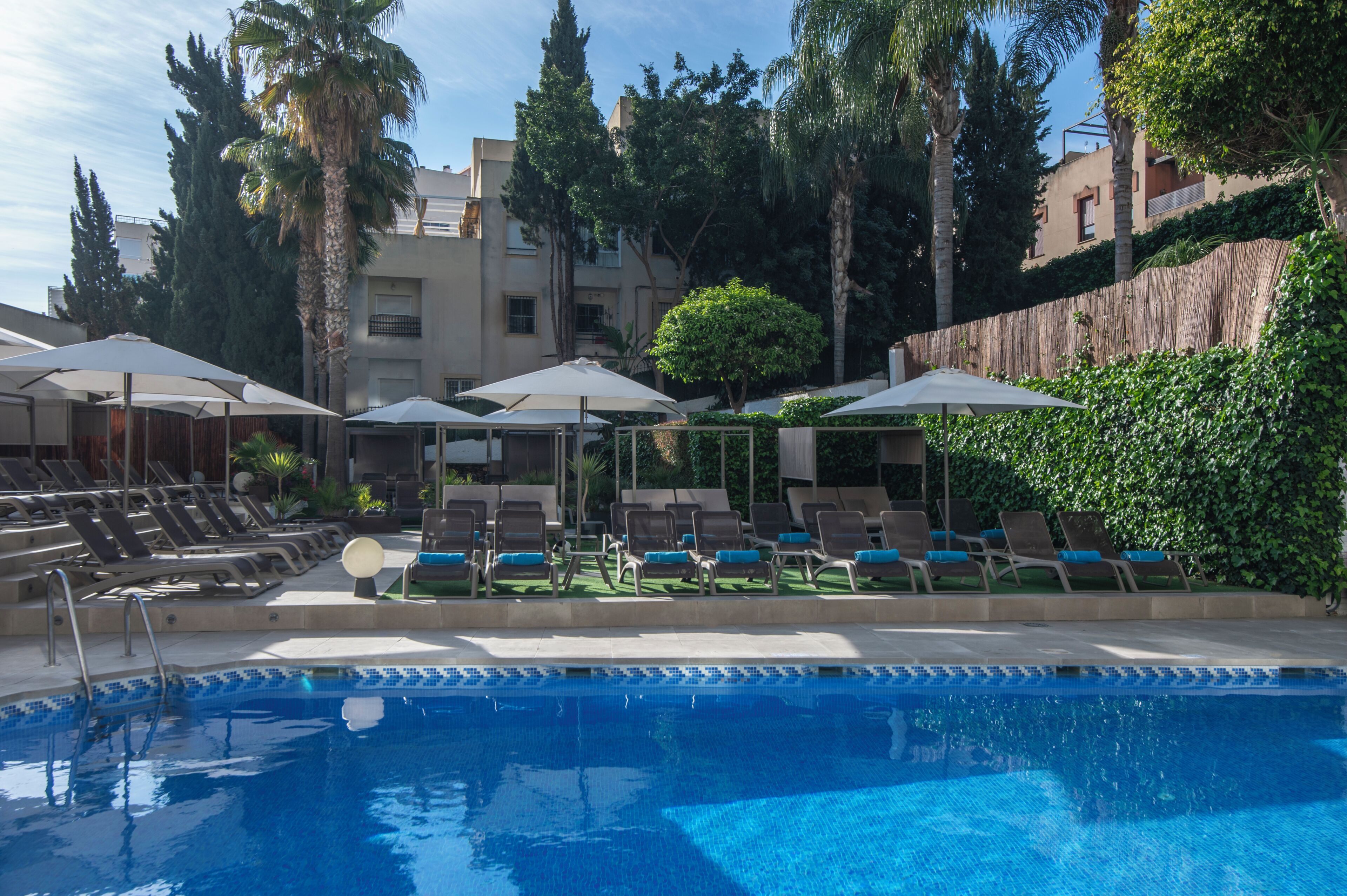 A swimming pool with lounge chairs and umbrellas, surrounded by greenery and buildings.