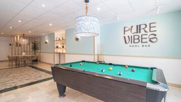 Une table de billard avec des billes, une queue de billard et un panneau indiquant « PURE VIBES POOL BAR », dans une pièce dont le sol est carrelé et qui est éclairée par un luminaire suspendu.