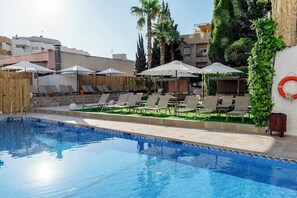 Une piscine entourée de verdure et de bâtiments, avec des chaises longues et des parasols.