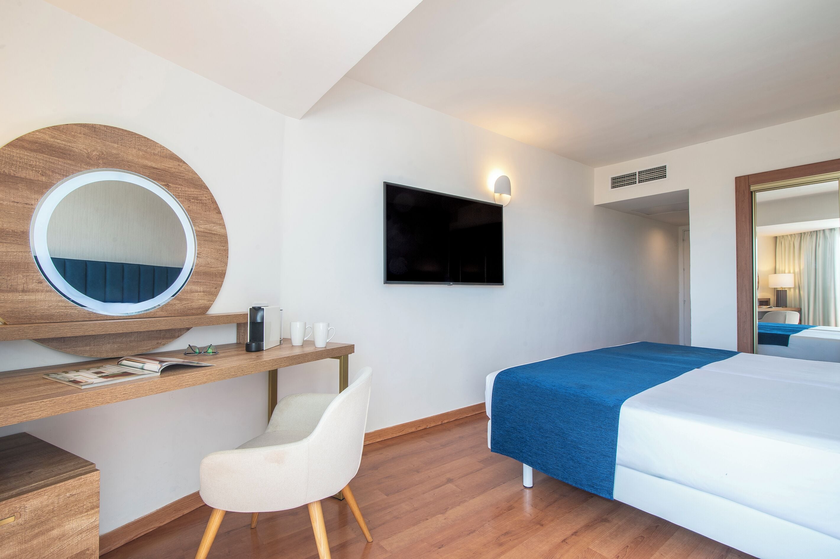 Habitació d'hotel amb escriptori de fusta, un mirall rodó, televisió de pantalla plana, llit amb edredó blau i cadira blanca.