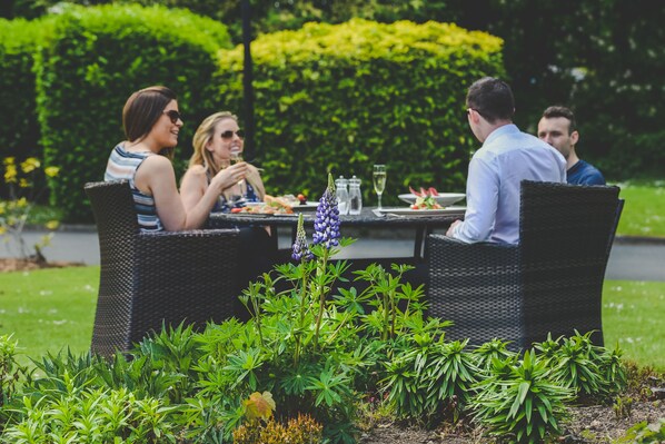 A group of people dining at an outdoor table with wicker chairs, surrounded by lush greenery and a well-maintained garden.