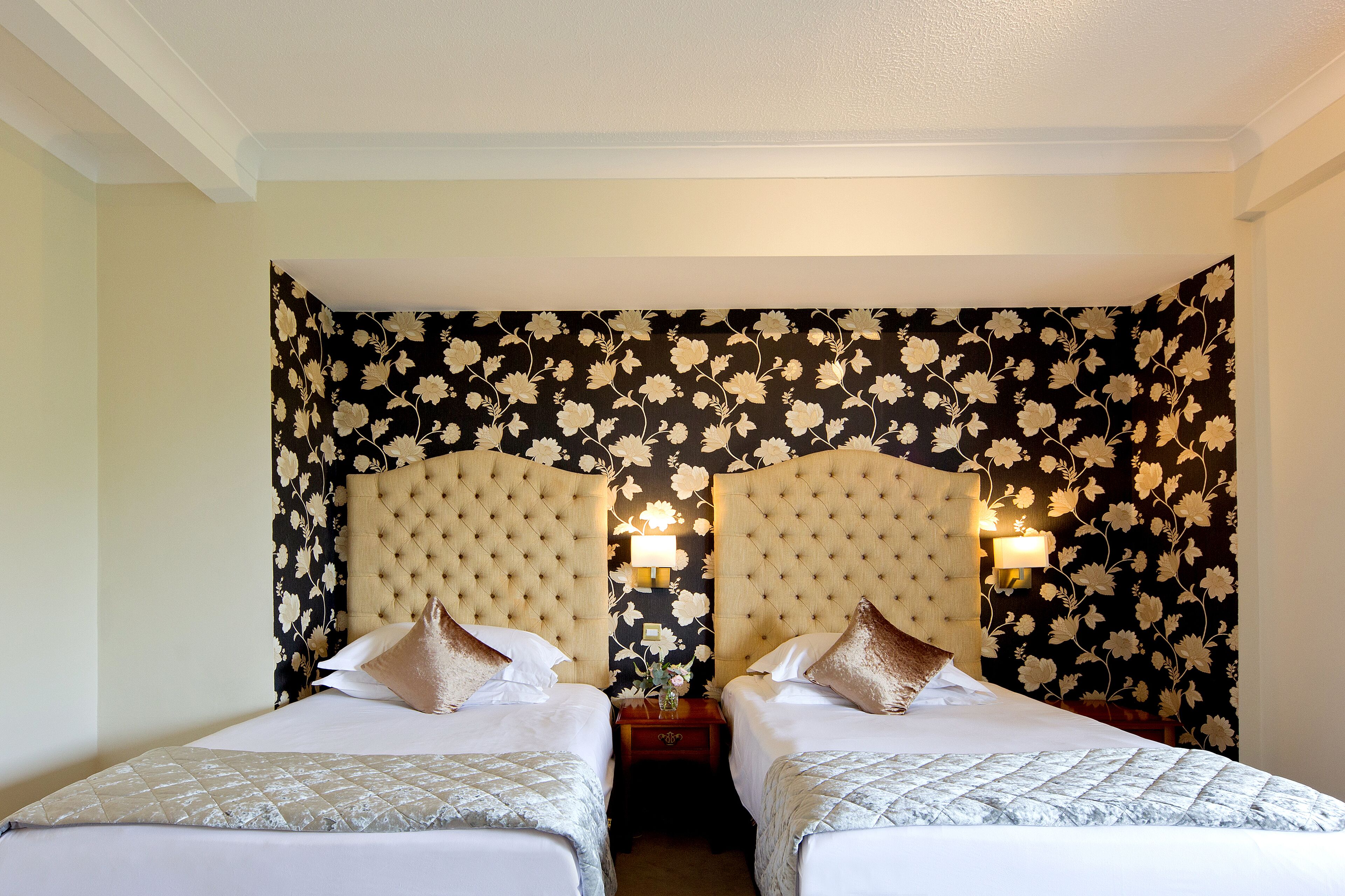 Habitación de hotel con dos camas, un diseño de papel tapiz floral y lámparas fijadas a la pared.