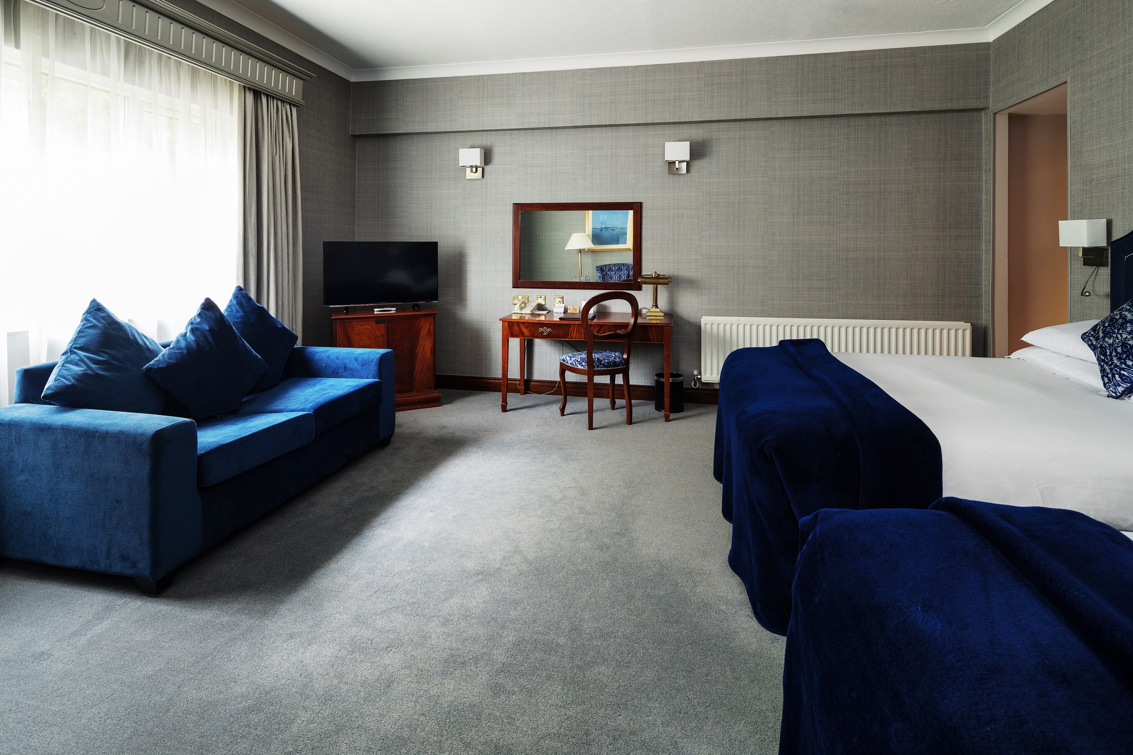 A hotel room with a bed, a sofa, a desk, a chair, a television, and a window with curtains.