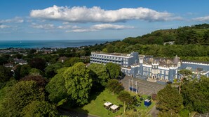 A large, historic building with multiple floors and a prominent tower, surrounded by lush greenery and a view of the sea.