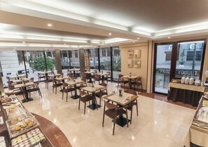 Un'area ristorazione moderna con diversi tavoli e sedie, un buffet con varie pietanze e ampie finestre che offrono vista sulla città.