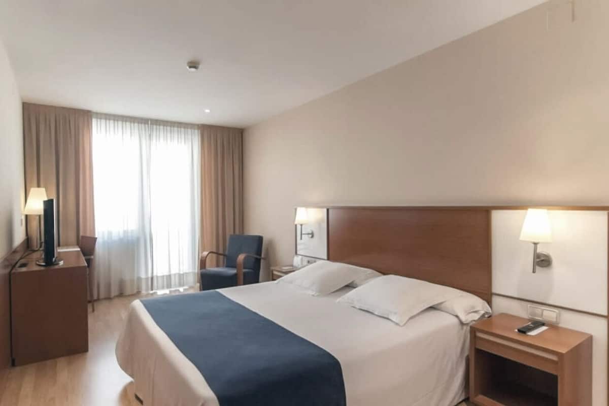Quarto de hotel com uma cama grande, duas mesas de cabeceira, uma cadeira e uma escrivaninha com abajur.