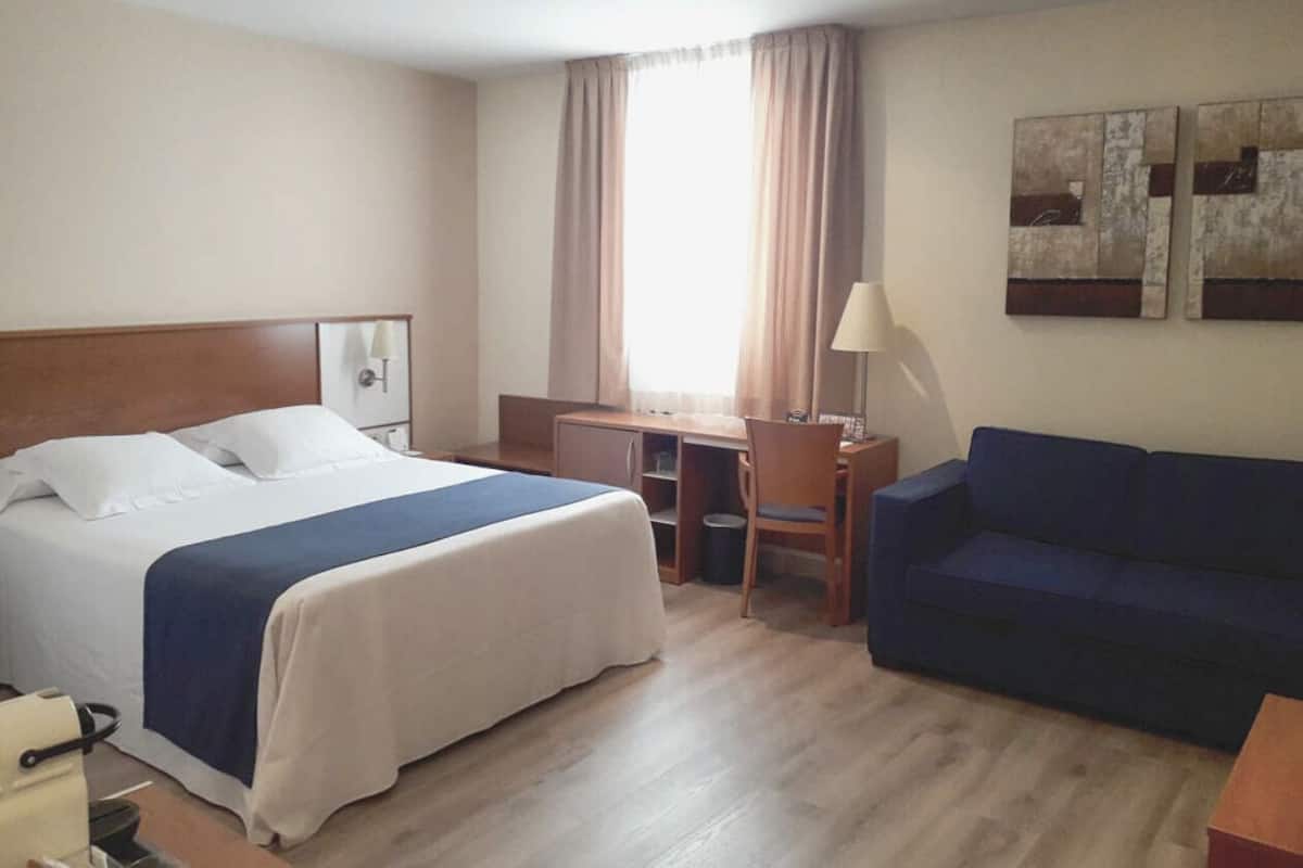 Quarto de hotel com cama, escrivaninha, cadeira, sofá e duas pinturas na parede.