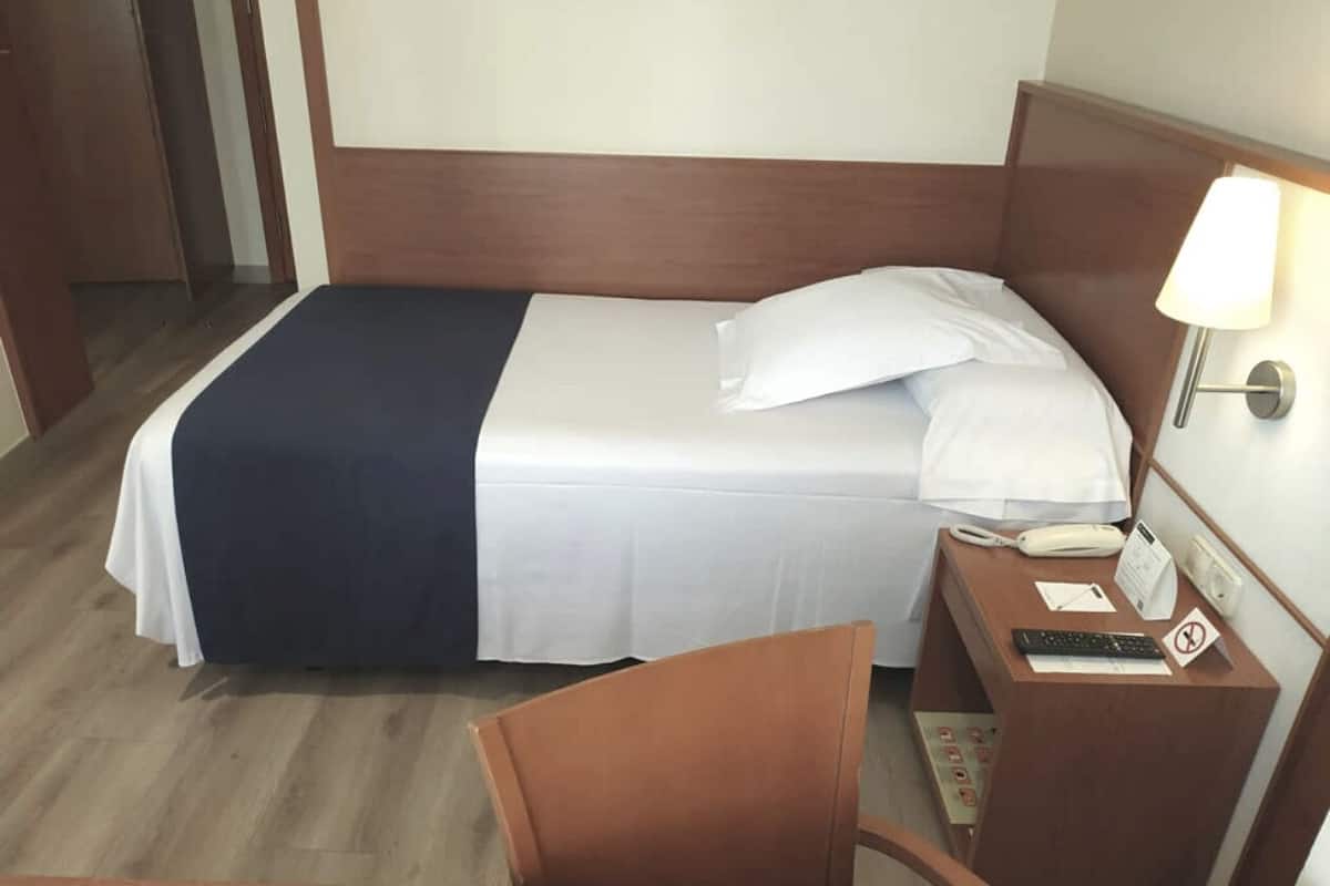 Quarto de hotel com uma cama, uma escrivaninha com telefone, uma cadeira e uma mesa de cabeceira com abajur.