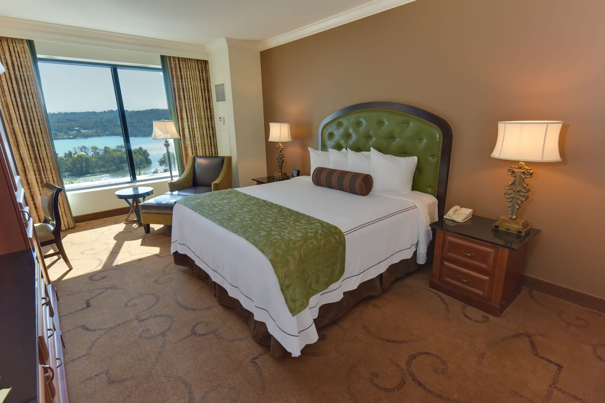 Habitación de hotel con una cama grande, dos mesitas de noche, una silla y vistas a un lago.