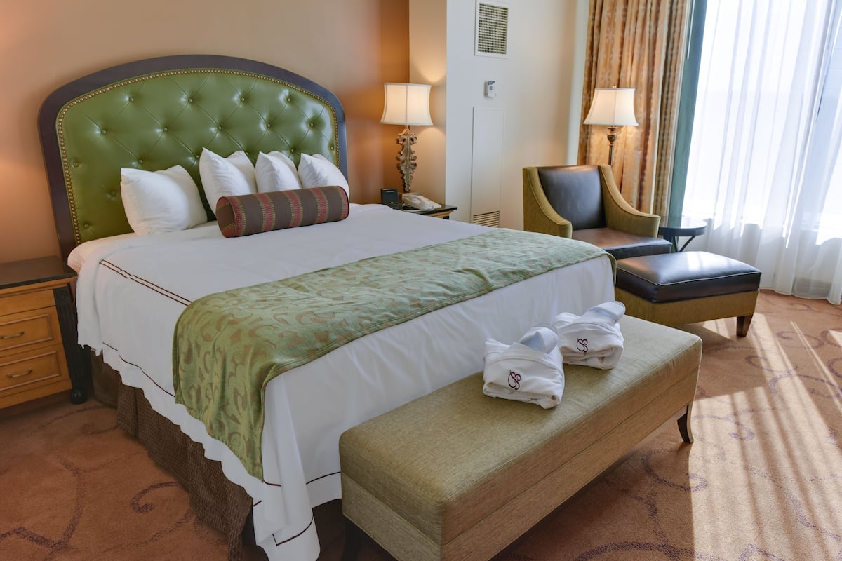 Habitación de hotel con una cama grande, un área de descanso con una silla y un reposapiés, y una mesita de noche con lámpara.