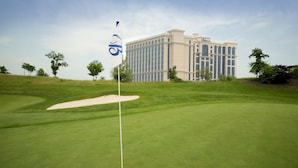 Ein Golfplatz mit Fahne, Sandbunker und einem Gebäude im Hintergrund.