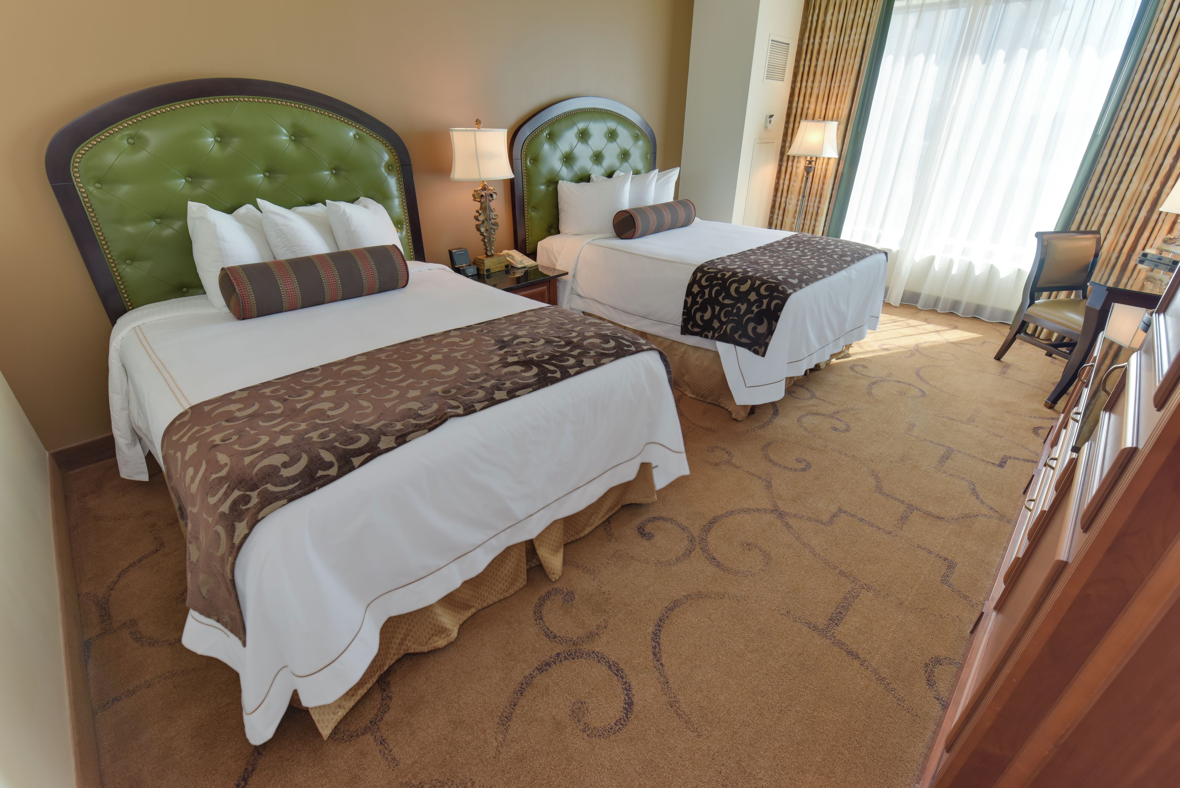 Une chambre d’hôtel avec deux lits, chacun doté d’une tête de lit verte, de draps blancs et d’un plaid à motifs marron. On y trouve une table de chevet avec une lampe, un téléphone et une petite boîte. La chambre est recouverte d’une moquette à motifs circulaires et possède une fenêtre avec des rideaux.