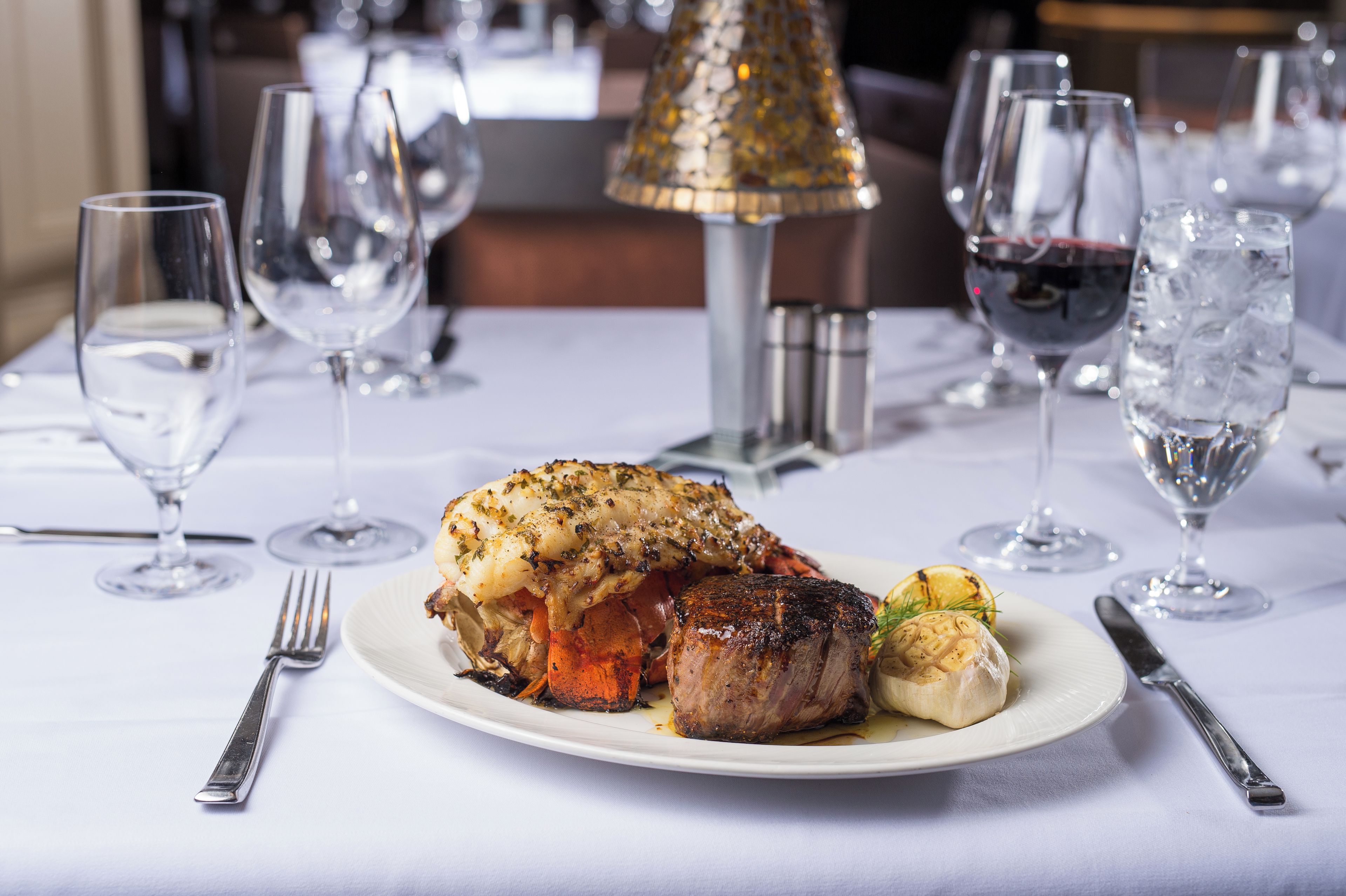 A plated meal with a lobster tail, a piece of meat, and a vegetable, accompanied by a fork and knife on a table with wine glasses and a decorative lamp.