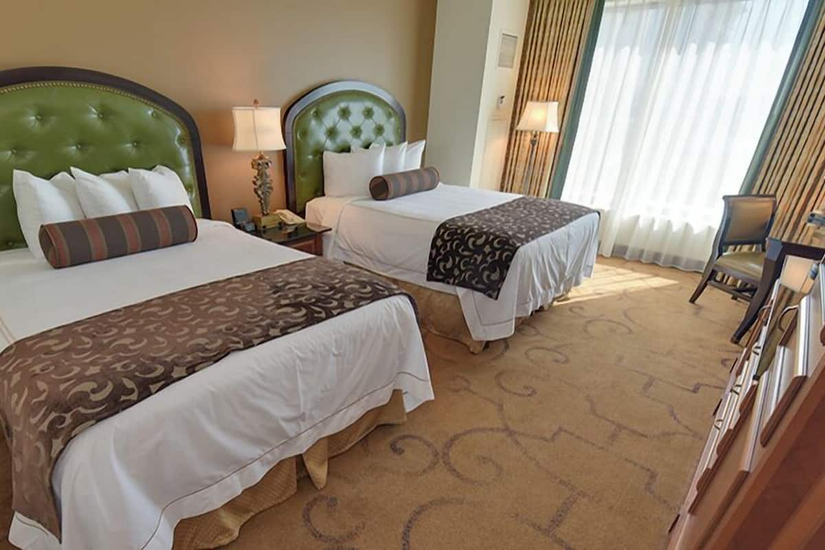 Habitación de hotel con dos camas, cada una con cabecero verde, ropa blanca y una manta estampada. Hay una silla y una lámpara sobre una mesita de noche.