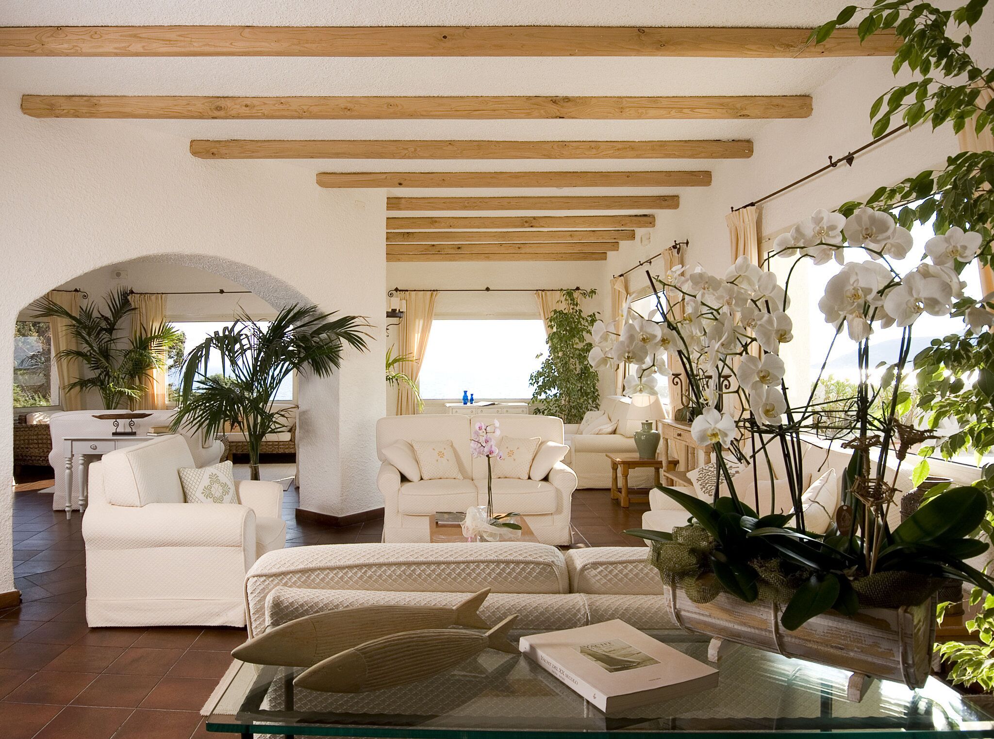 A spacious living area with a high ceiling, wooden beams, and a view of the sea. There are multiple seating areas, including a sofa and armchairs, and a dining table with chairs. The room is decorated with white furniture and various plants, including orchids.