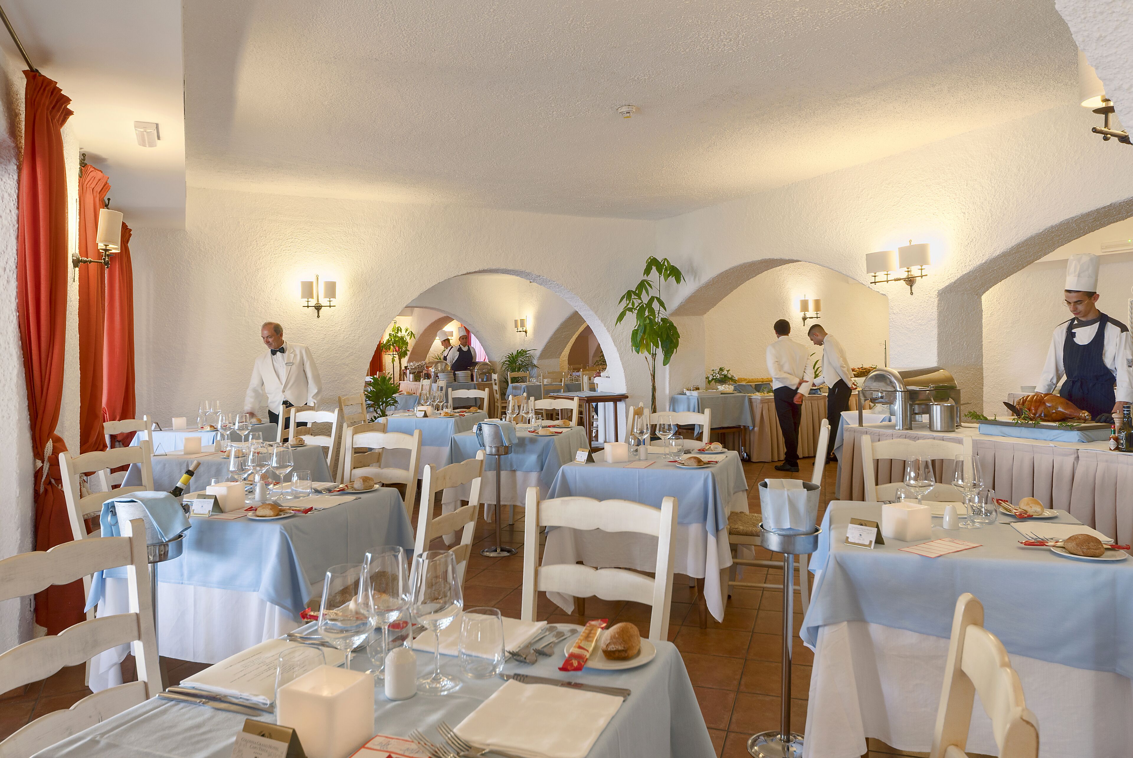 A restaurant with multiple tables set for dining, staff in uniform, and a chef preparing food.
