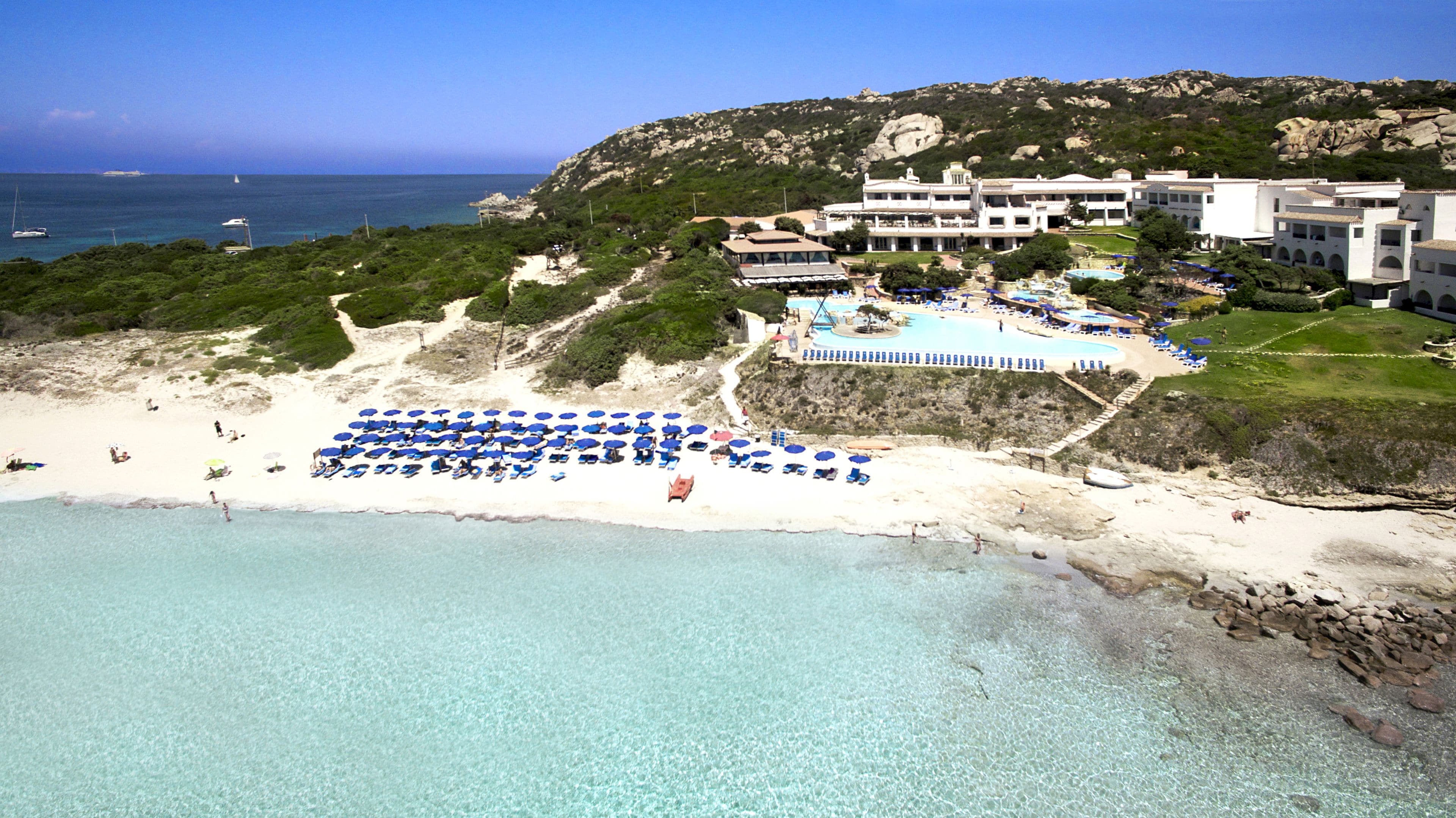 A coastal resort with a beach, a pool area, and a mountain backdrop.