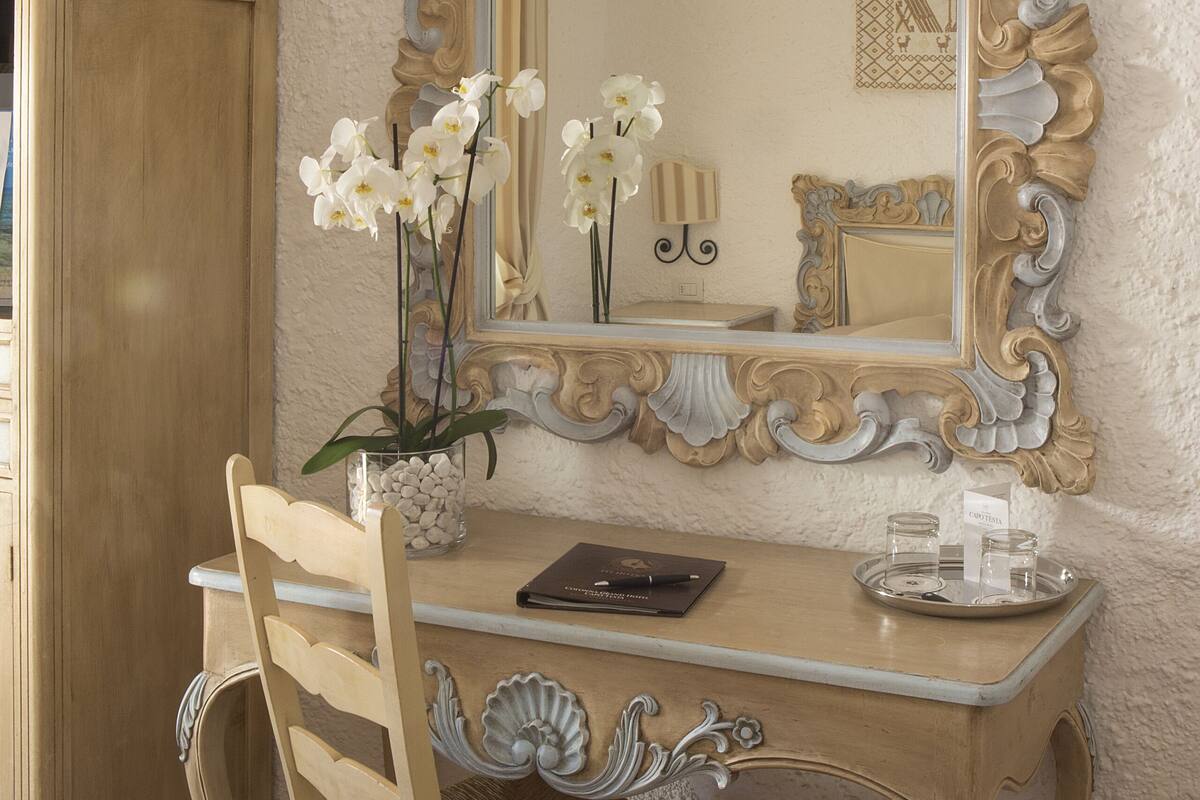 Une pièce comprenant un bureau en bois, une chaise, un miroir doté d’un cadre orné et un vase contenant des fleurs blanches.