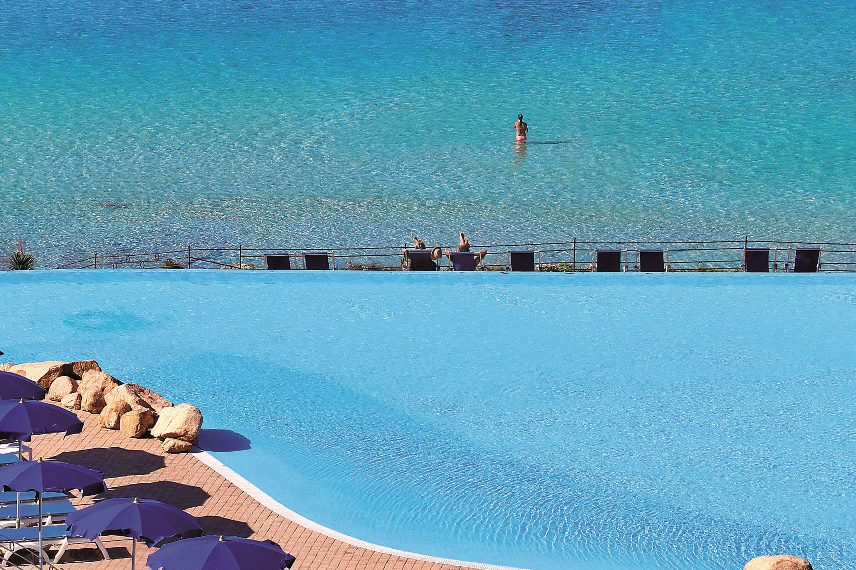 Une piscine d’un bleu éclatant, dotée de chaises longues et de parasols, un espace aménagé au bord de la piscine avec des rochers, et une vue sur la mer.