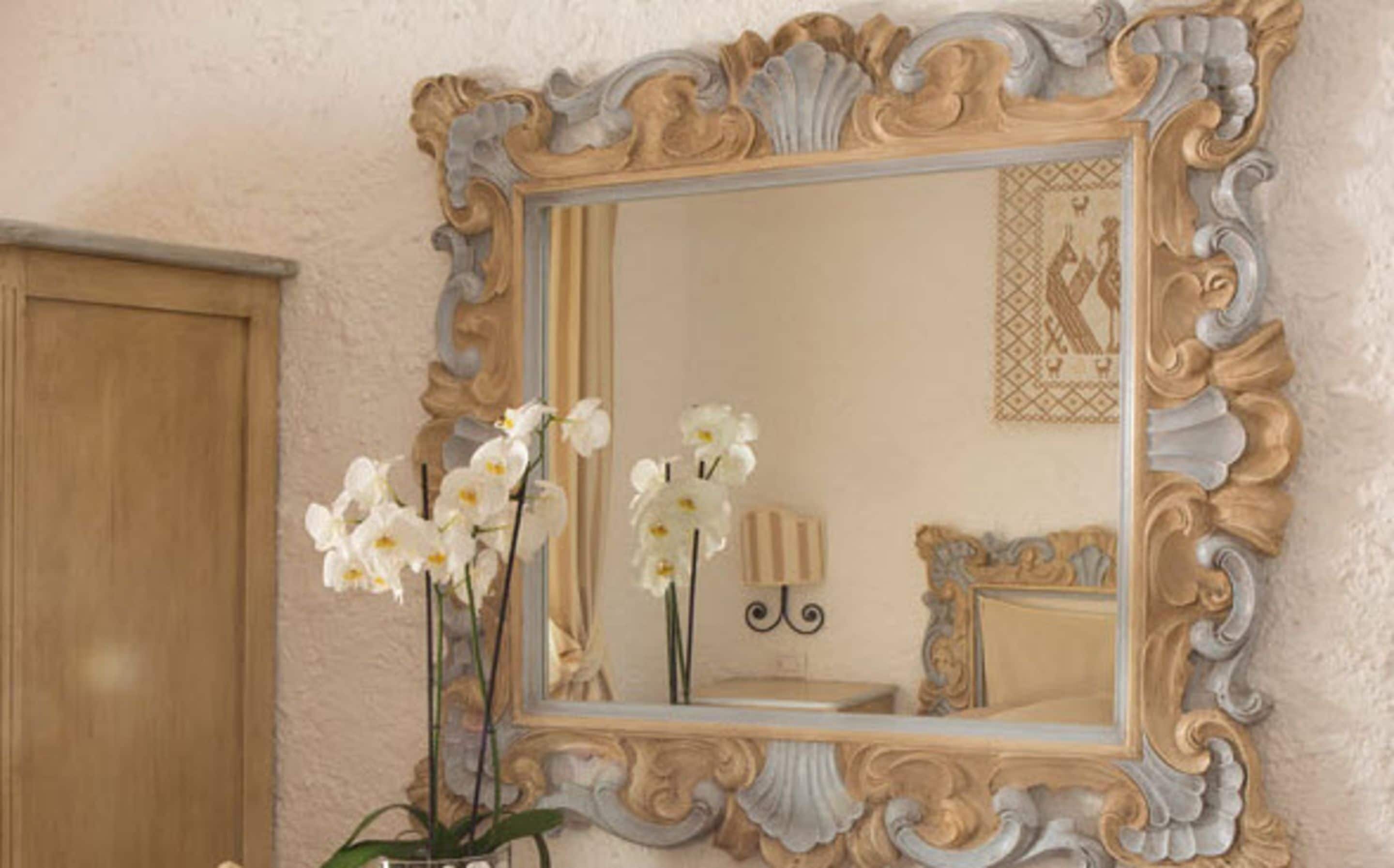 Un espejo con un marco dorado y blanco ornamentado, que refleja una habitación con una puerta de madera y un cuadro enmarcado en la pared.