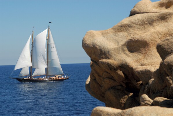 Un velero con varias velas en el agua, y una costa rocosa al frente.