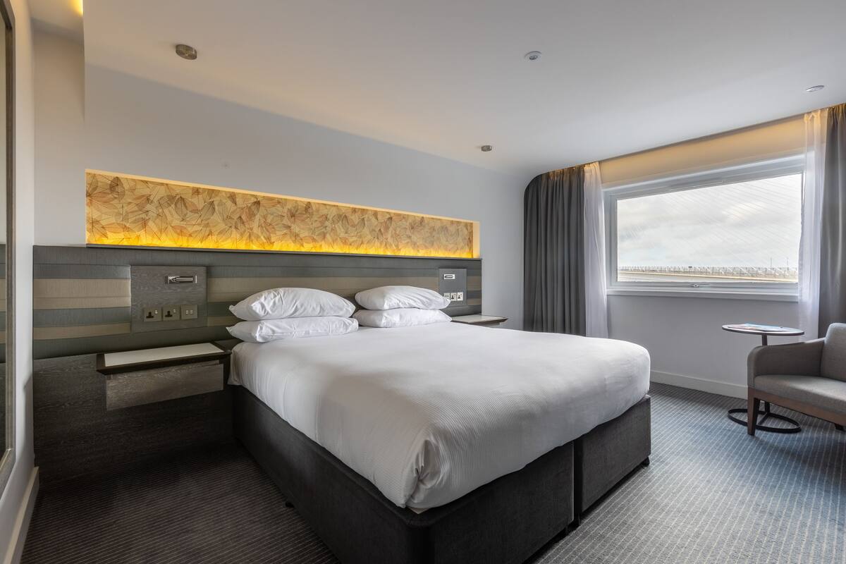 A modern hotel room with a large bed, a bedside table, a small desk, a chair, and a large window with a view.
