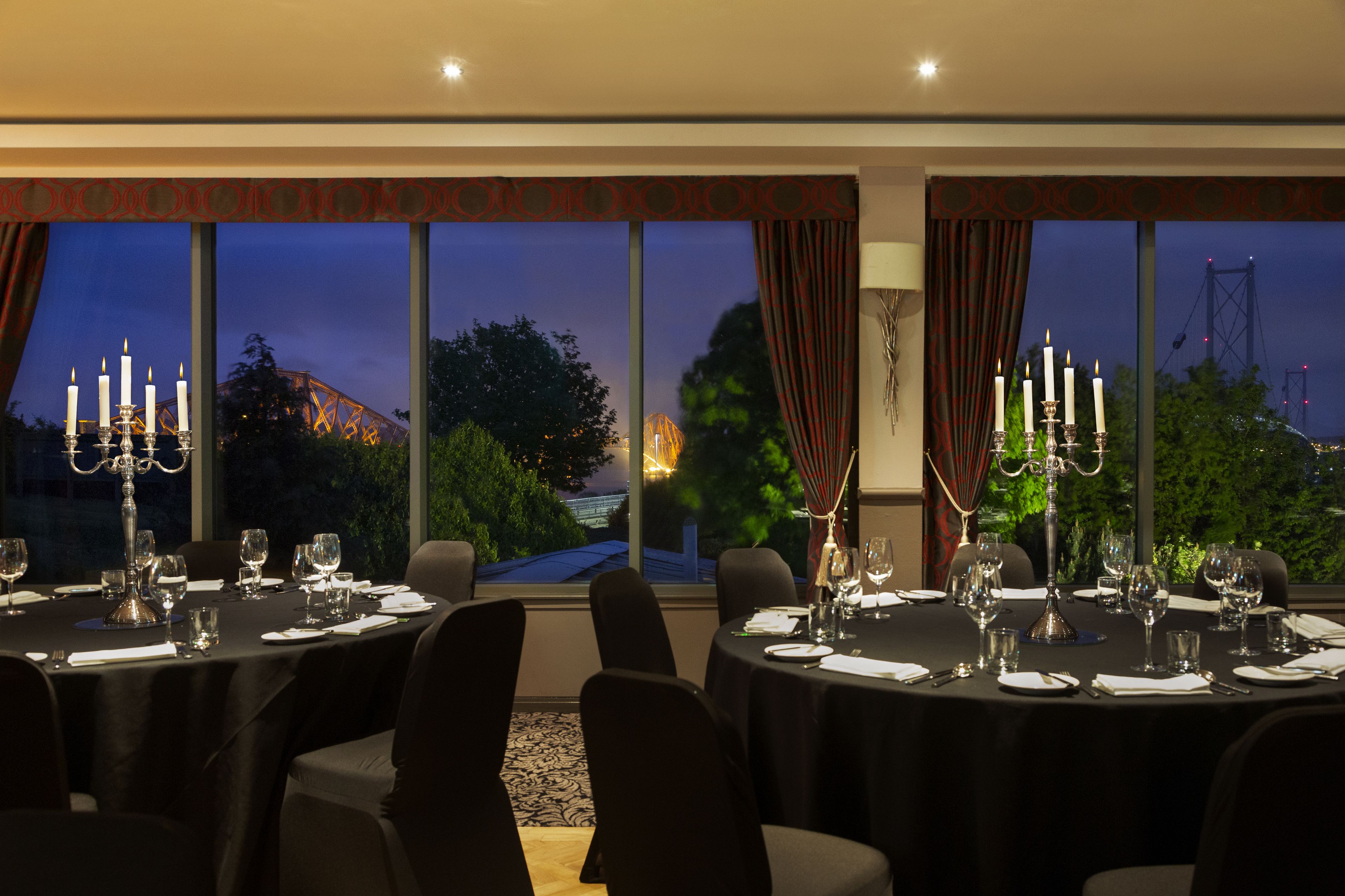 A dining room with tables set for a meal, large windows with curtains, and a view of a bridge at dusk.
