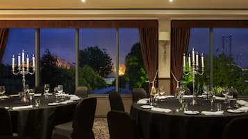 A dining room with tables set for a meal, large windows with curtains, and a view of a bridge at dusk.