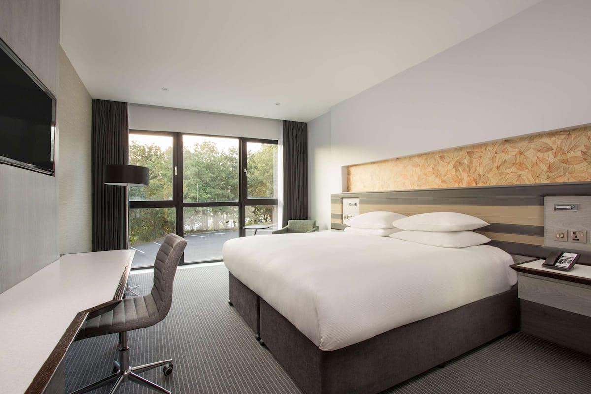 A hotel room with a bed, a desk, a chair, a TV, and a window with a view of trees.