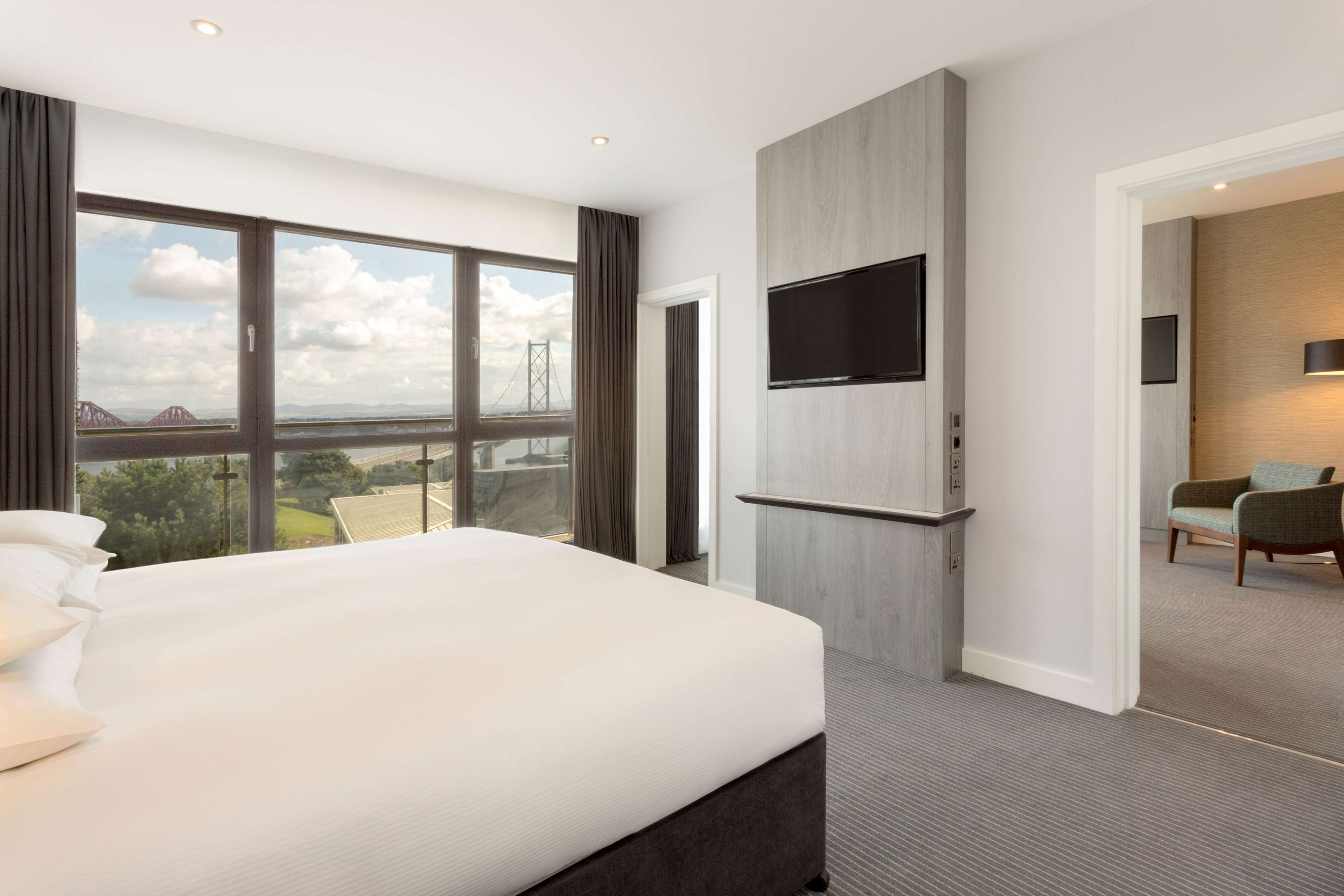 A modern hotel room with a large bed, a TV mounted on the wall, and a view of a bridge and landscape through large windows.
