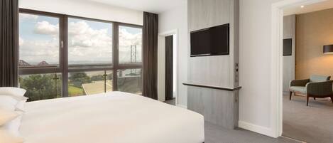 A modern hotel room with a large bed, a TV mounted on the wall, and a view of a bridge and landscape through large windows.
