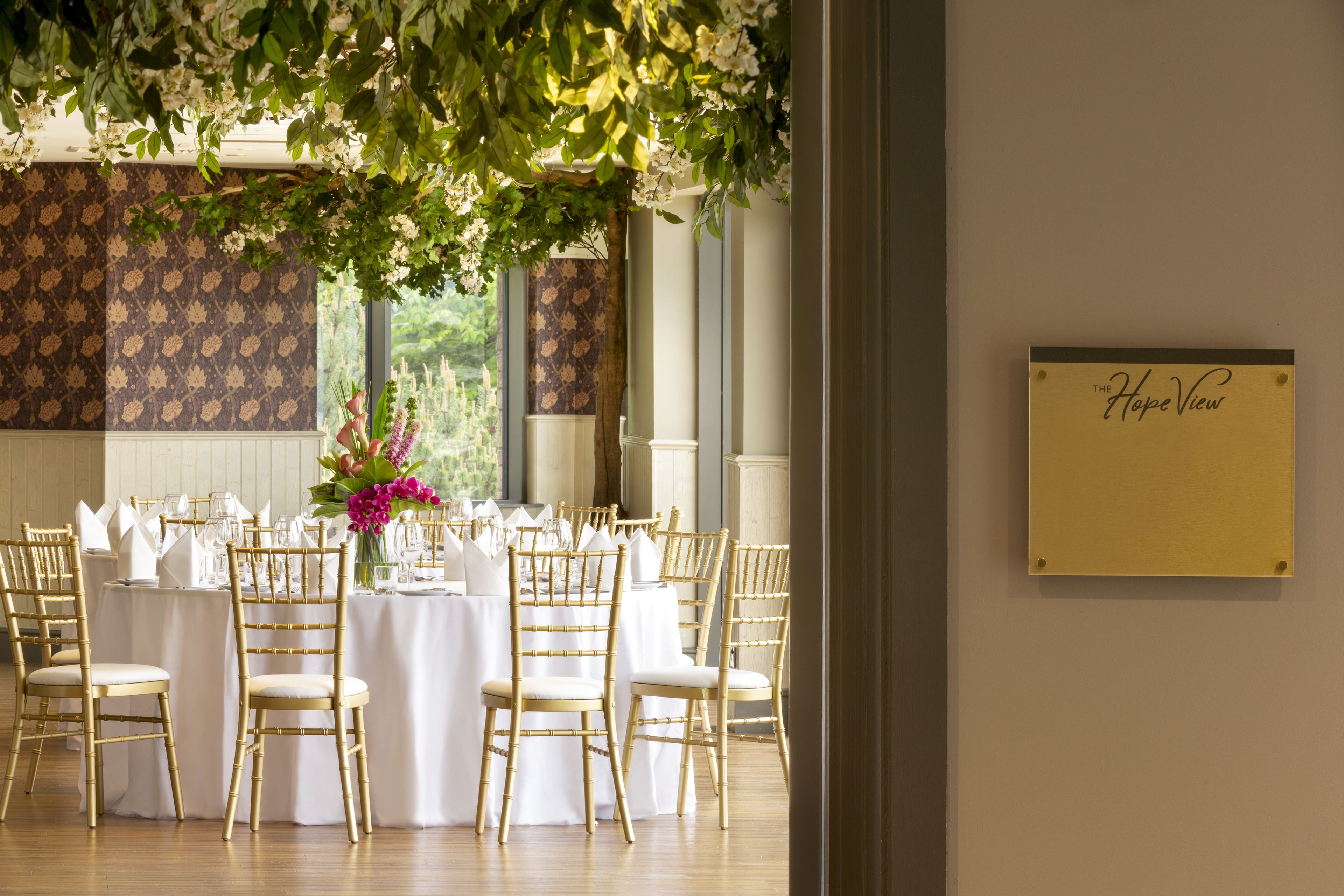 Un salón de banquetes con mesas redondas dispuestas para un evento formal, con manteles blancos y sillas doradas.