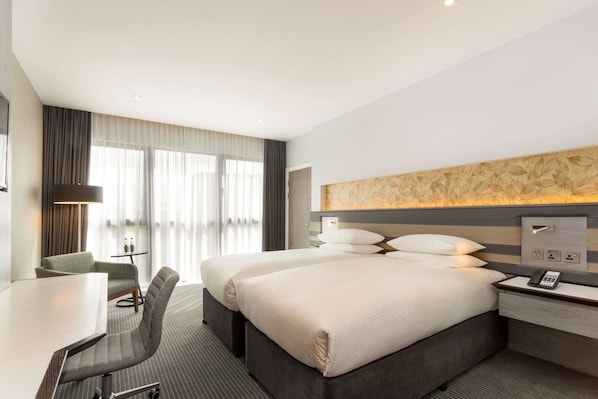 A hotel room with two beds, a desk, a chair, a small table, and a large window with curtains.