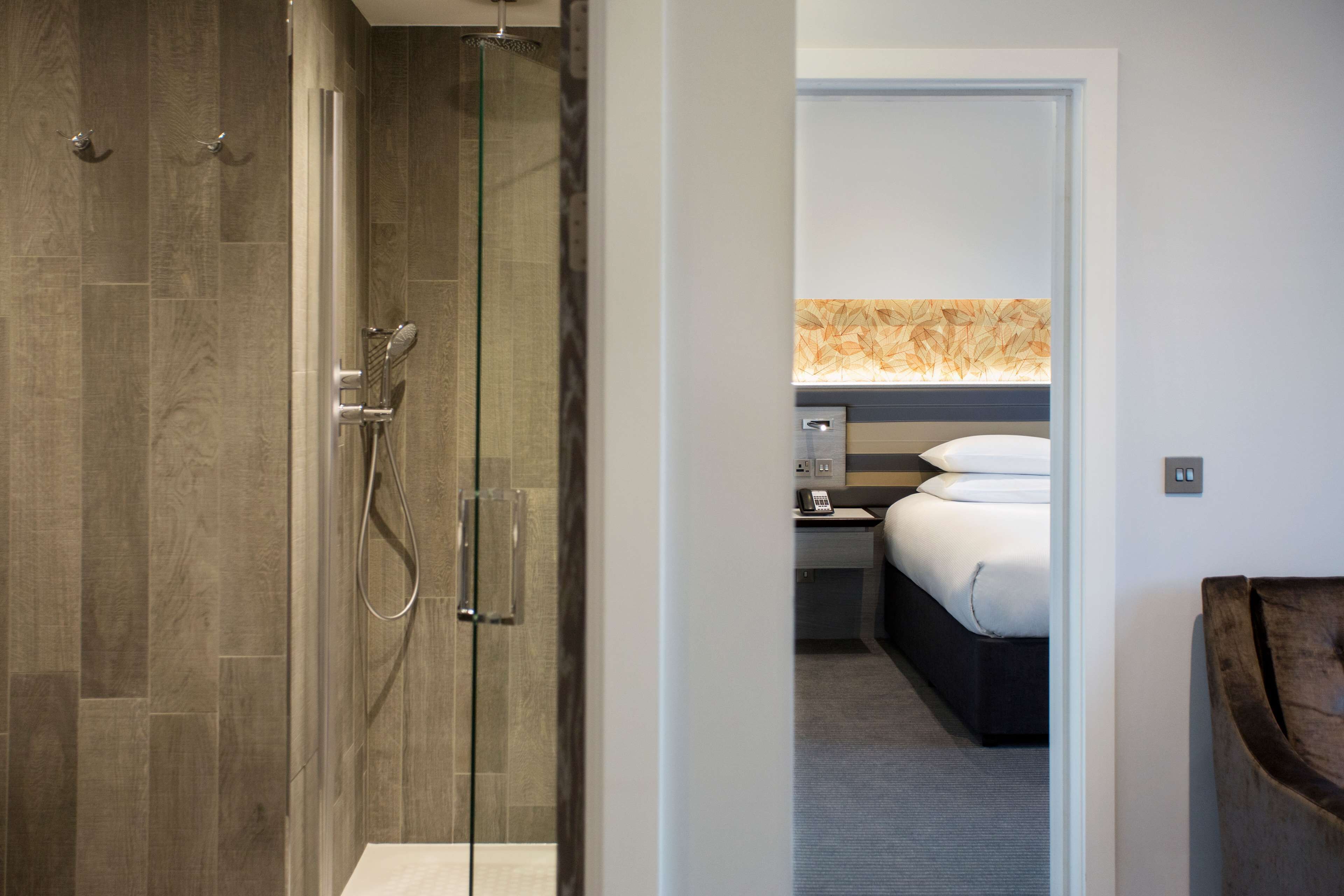 A modern hotel room with a glass shower enclosure, a bed with white linens, and a nightstand with a telephone.