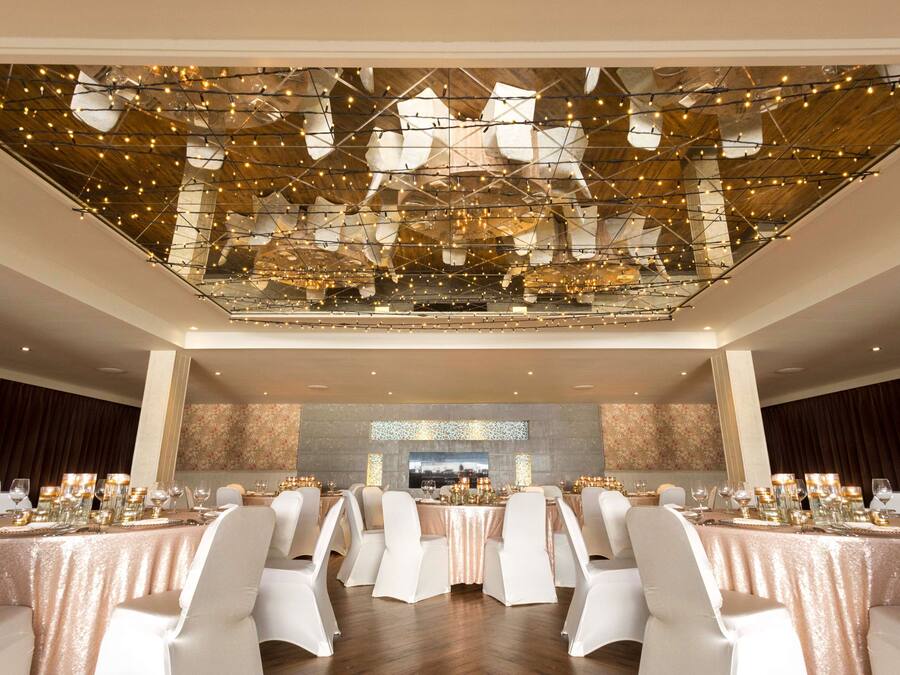 A banquet hall with tables set for a formal event, featuring a high ceiling with a geometric light fixture and a large wall-mounted screen.