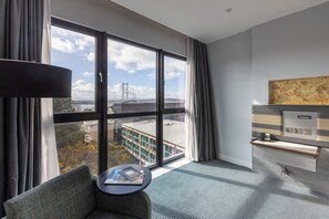 A modern hotel room with a large window, a sofa, a small table with books, and a desk with a computer.