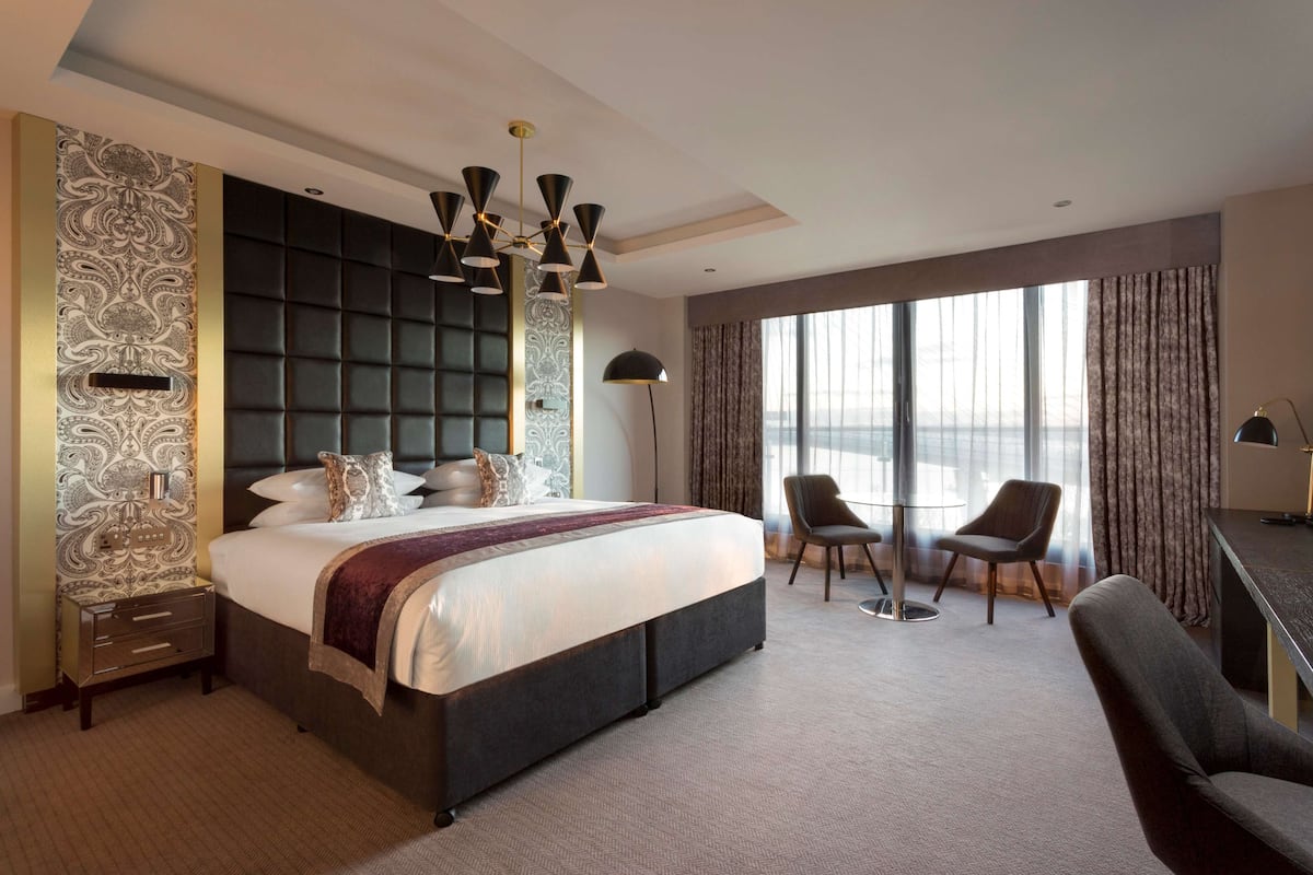 A modern hotel room with a large bed, two chairs, a desk, and a view of the city.
