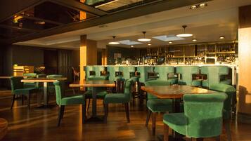 A restaurant with green upholstered chairs and wooden tables, a bar area with shelves of bottles, and a high ceiling with recessed lighting.