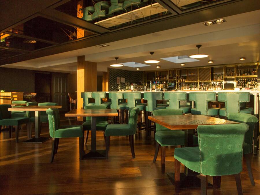 A restaurant with green upholstered chairs and wooden tables, a bar area with shelves of bottles, and a high ceiling with recessed lighting.