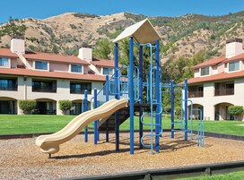 Un parque infantil con tobogán y estructuras para escalar, con edificios residenciales y techos de tejas rojas al fondo.
